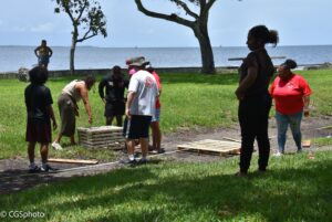 A group of people working together outside. in the background is the ocean. The group is standing around some wooden pallets looking together, other people are in the foreground and background looking at the group. A woman wearing a sleeveless grey top, dark green pants, and sneakers points at something on the pallet on the left, while others stand around looking at what she is doing. On the far right, two women stand looking at the other figures. The woman on the right wears a red polo shirt with the DWP logo on it, and blue jeans. Her hair is tied back into a bun. The woman standing closer to the viewer is in shadow, and wears a dark colored top and pants, and has her hair in a loose bun.