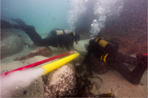 two divers laying on the ocean floor looking at something between them. they are surrounded by large rocks and a pipe spraying water and particles away from them.