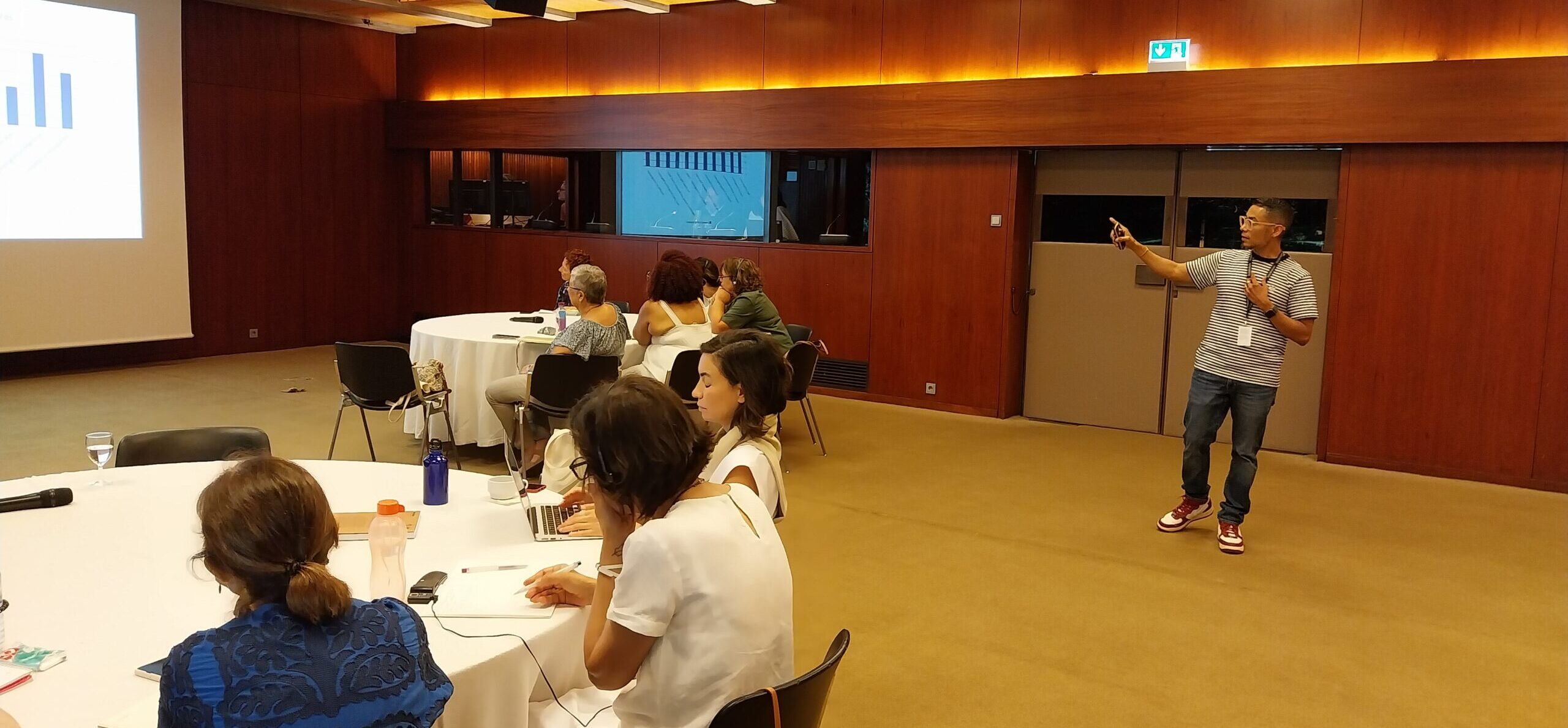 man standing in a room pointing a presentation screen while others take notes