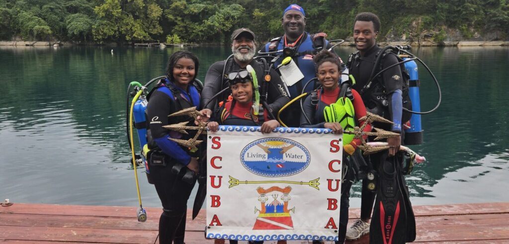 Six people standing on a dock in full scuba gear posing for a photo with a sign