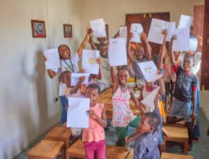 group of children hold up worksheet and smile at the camera in a classroom