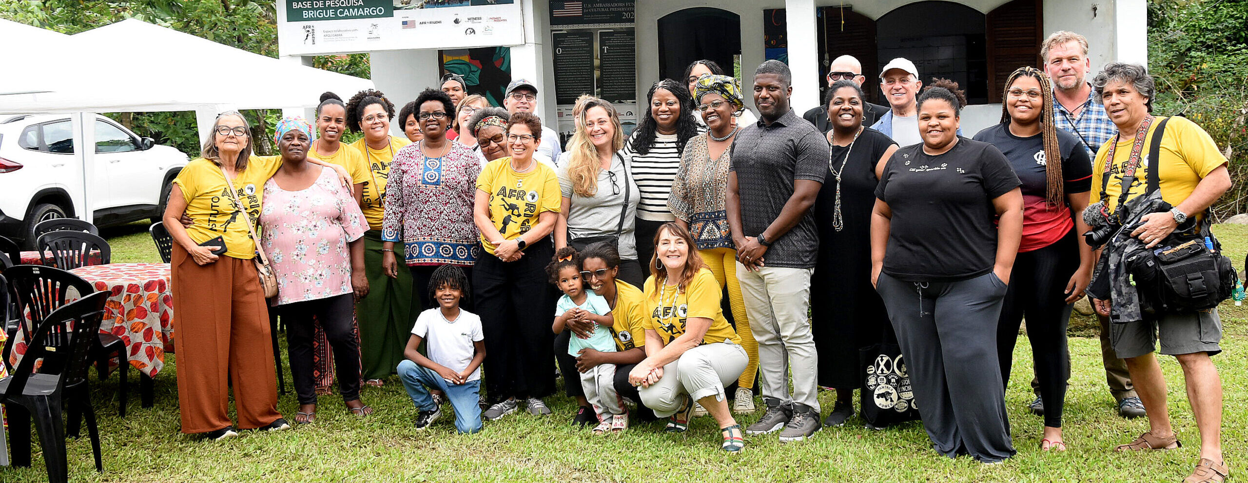 29 people pose for a photo on the grass in front of a small building reading "Base de Pesquisa Brigue Camargo.