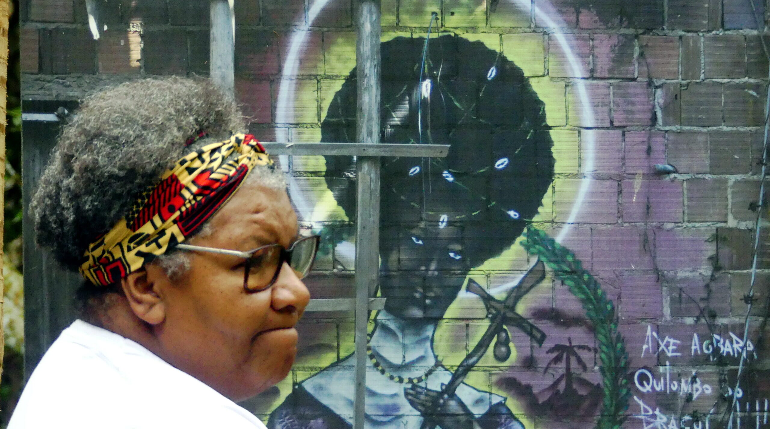 woman looks across the frame. Behind her a there is a painting on a saint on the brick side of the building looking at the camera.