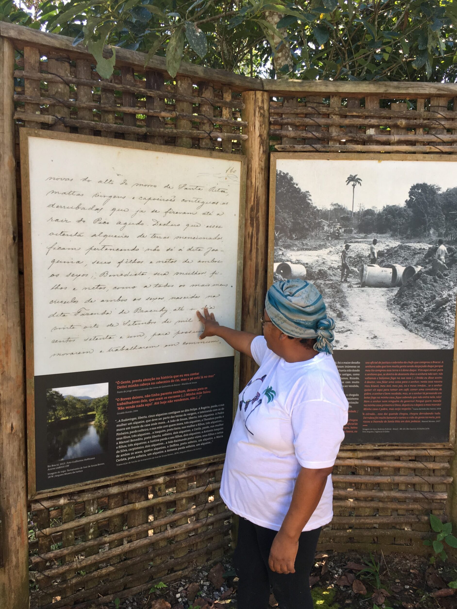 Woman faces towards exhibition panels pointing to a written document. Trees are visible in the background.