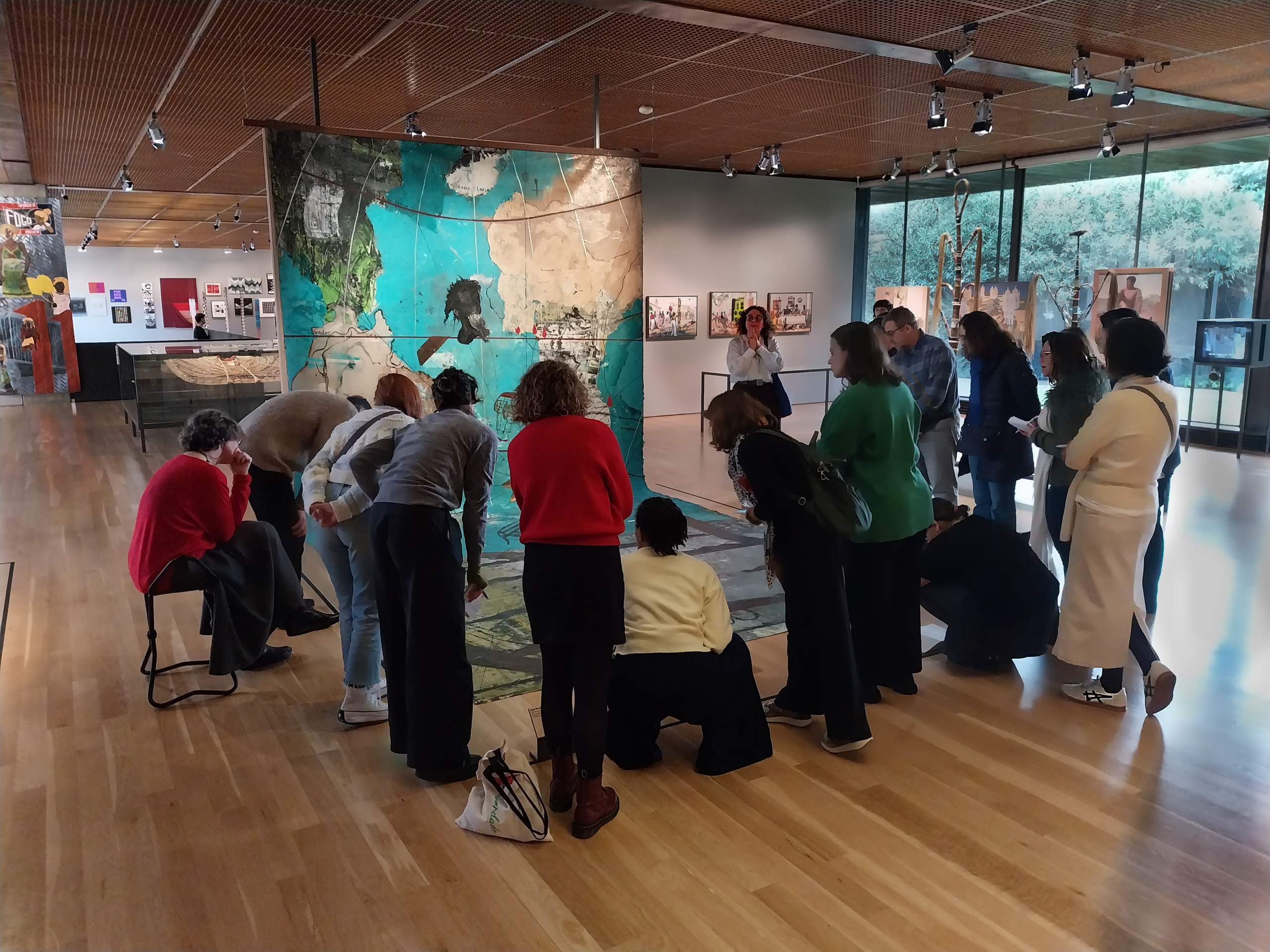 tour group looks at piece of art in the middle of a gallery. Everyone looks away from the camera and at the piece which includes a map of the atlantic world on the ground and suspended on a canvas