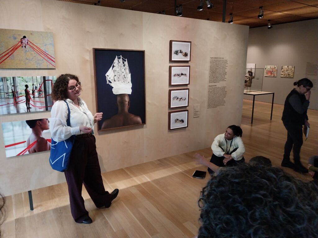 Woman standing in front of a a wall of art in a gallery. The central painting on the wall is of a black man facing away from the audience with a completely white ship balanced on his head
