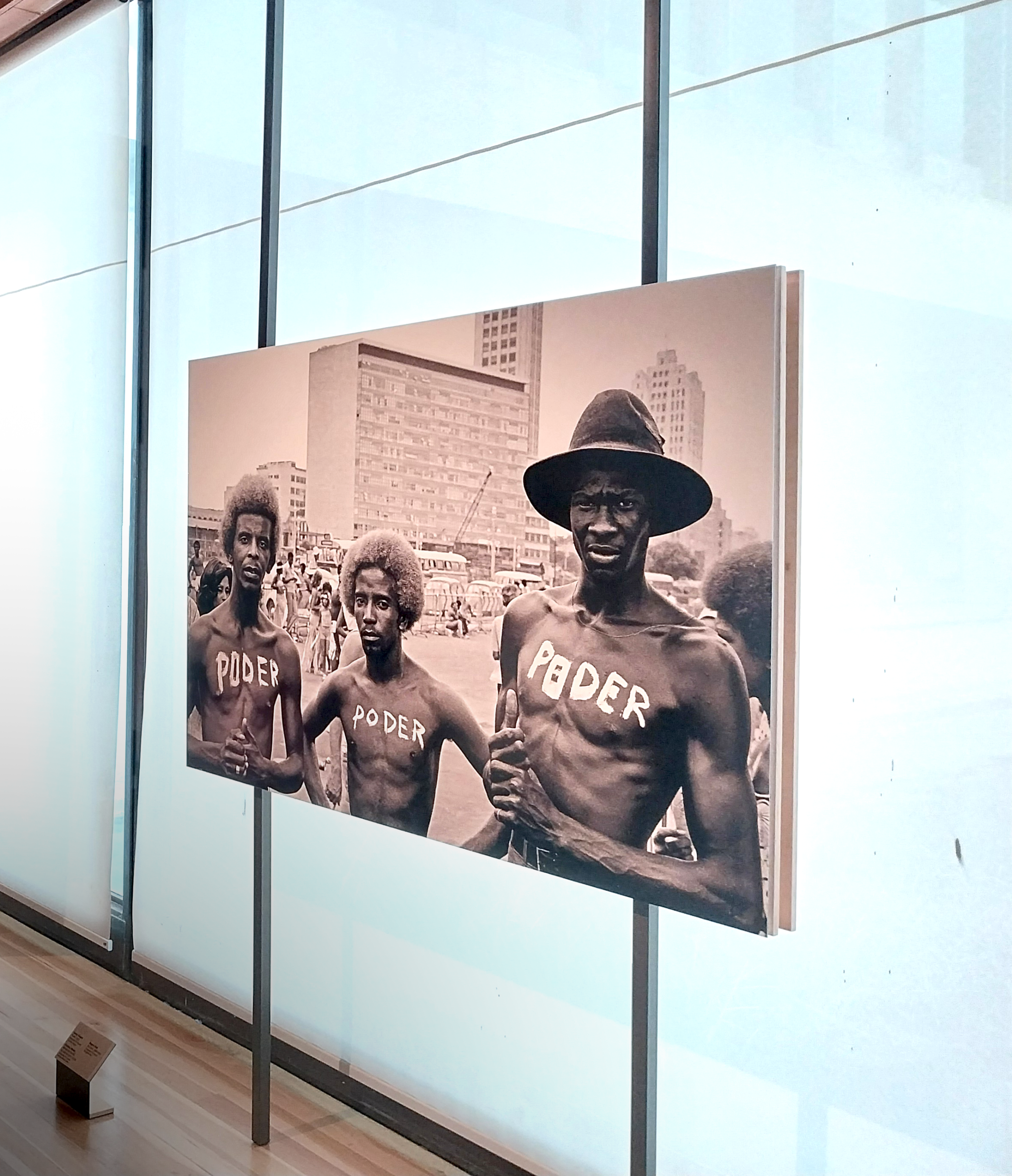 A photo displayed in trhe wall of the gallery. The image is black and white and shows three Black men shirtless with the word "PODER" written across their chests.