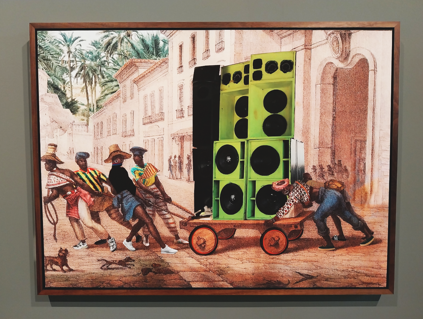 Photos shows a framed painting on a grey background. The painting shows four people pulling and one person pushing a cart down a historic street. The people, who are all Black are dressed in modern clothes. On the cart they carry 4 modern bright green speakers
