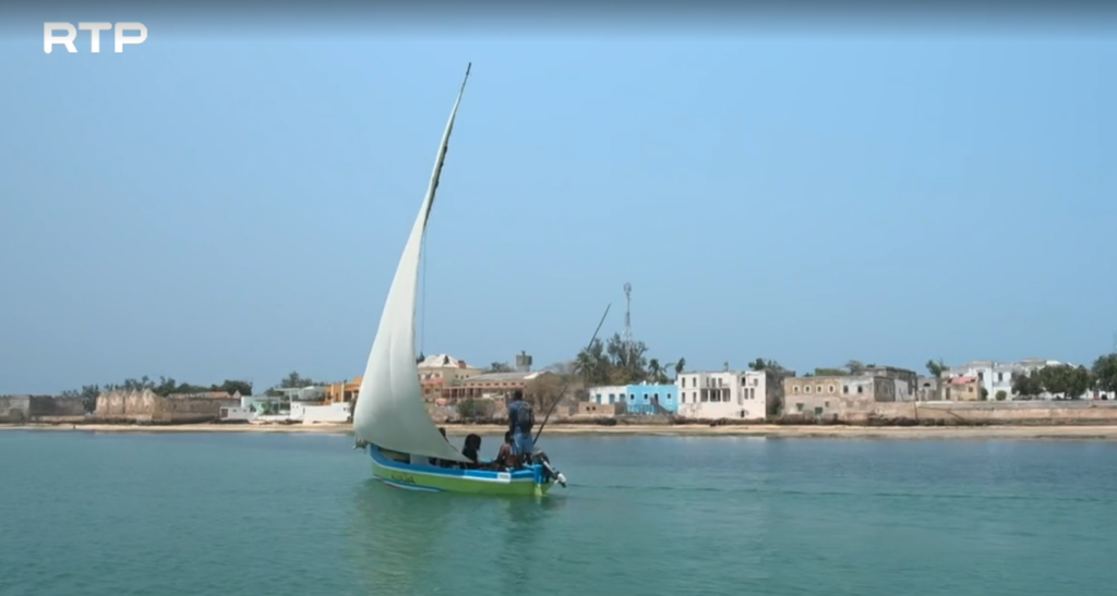 Sail boat is seen sailing near the coast on blue green water and a against a blue sky